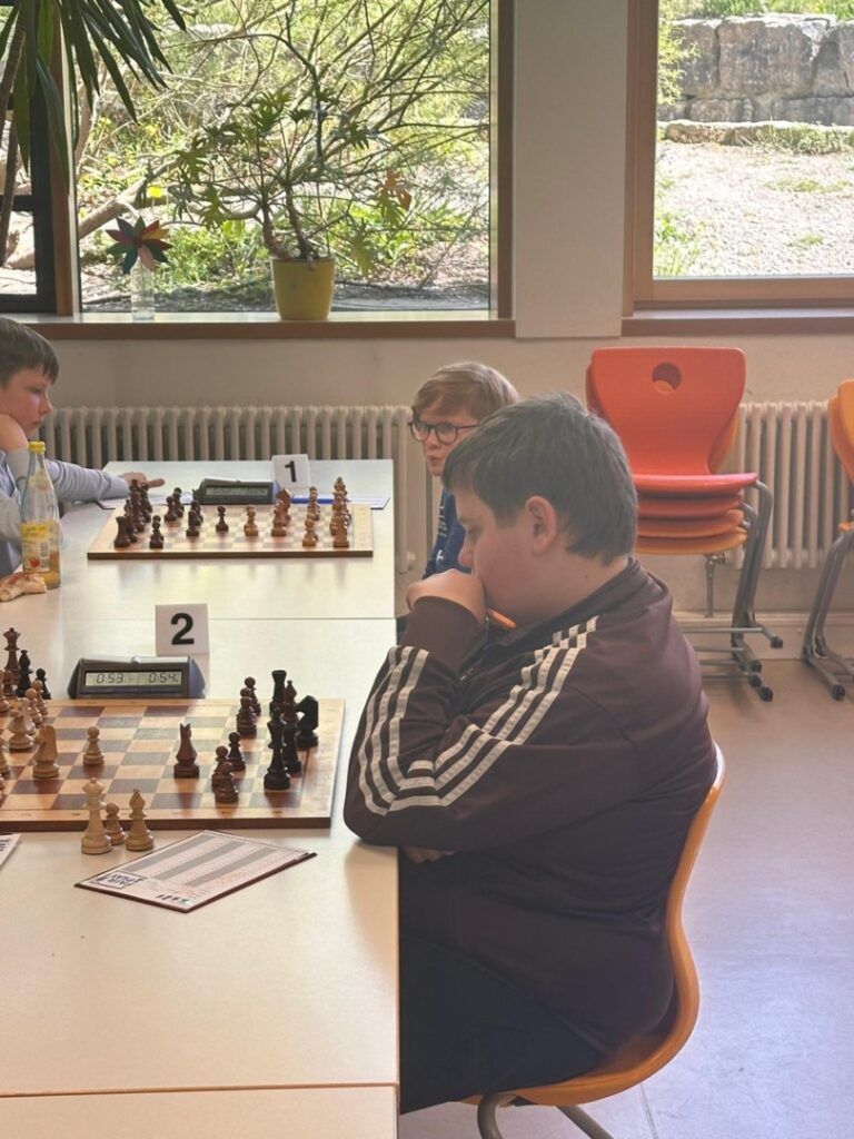 Boy in a brown jacket focuses on a chess match at a table with a clock and numbered board markers in a bright room.