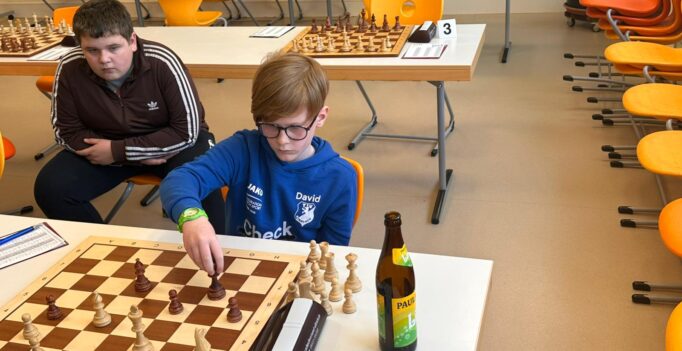 Two boys sit at a classroom chess table; the boy in a blue hoodie with glasses makes a move on a wooden chessboard while his opponent watches, with score sheets and a bottle on the table nearby.