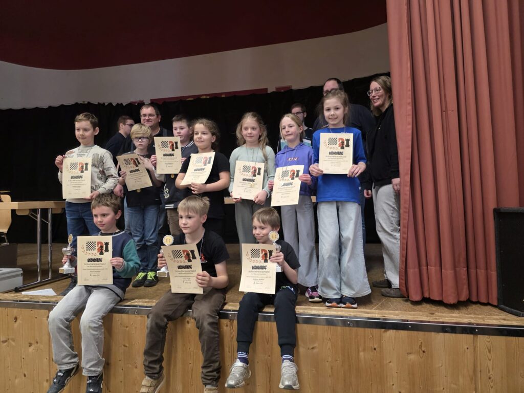 Group of children on a stage holding certificates after an awards ceremony, with a few adults in back and red stage curtains nearby.