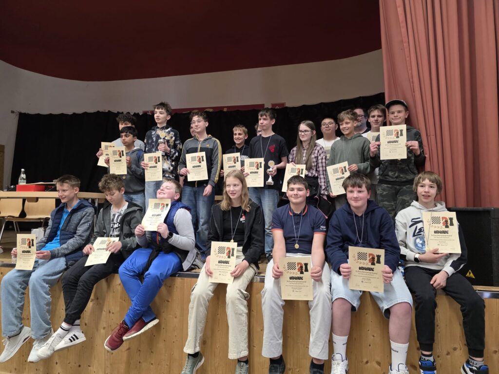 Group of young students on a stage holding certificates and medals after an awards ceremony in a school gymnasium.
