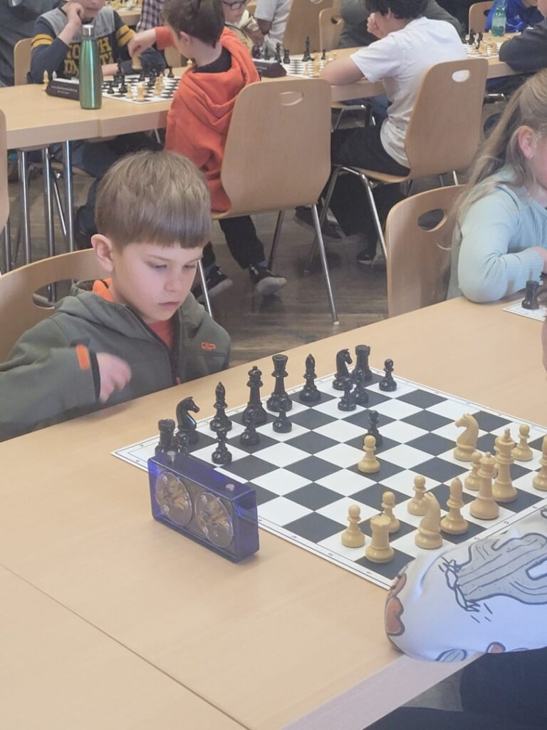 Young boy concentrates on a chess game at a classroom tournament, with a blue timer on the table and other players in the background.