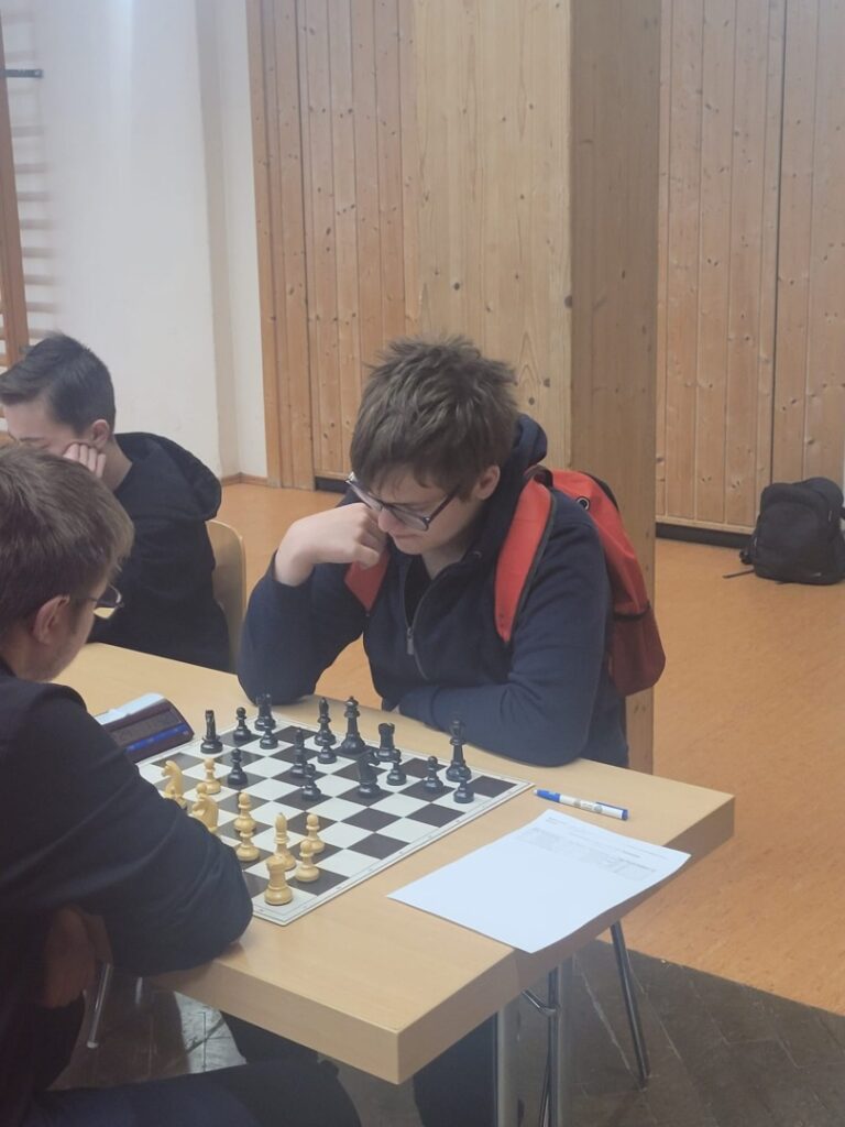 Two boys sit at a table playing chess; one wears a red backpack and glasses, studying the board intently.