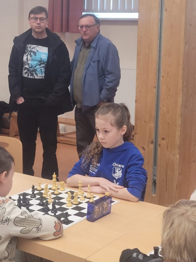Girl in a blue Chiara hoodie sits at a chessboard, concentrating on the game as two men stand behind her in a classroom or hall.