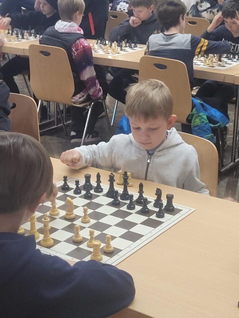 Children sit at tables playing chess in a classroom; a young boy in a gray hoodie focuses on the board at the foreground essaying a move.