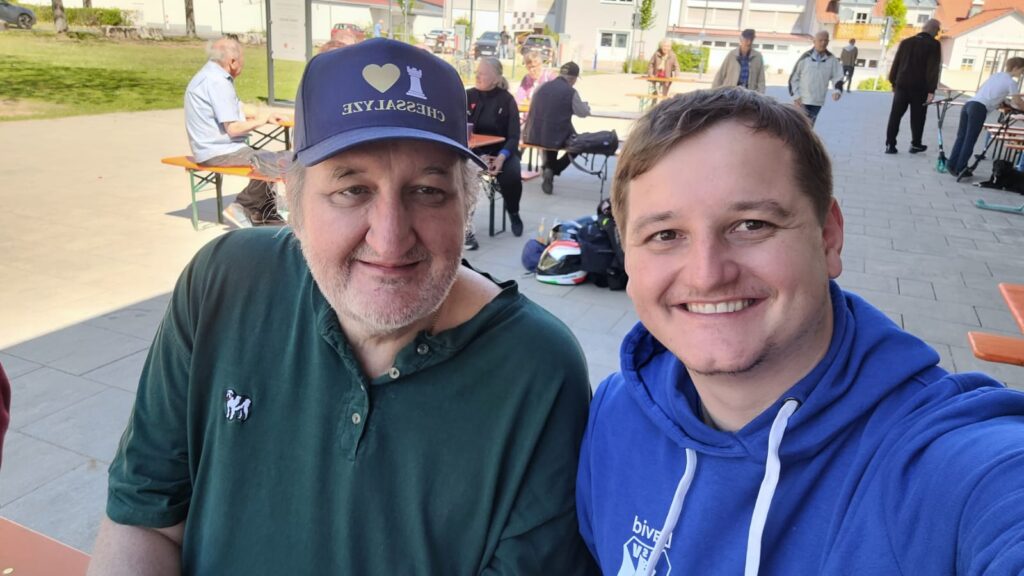 Two men taking a selfie at an outdoor community gathering: one in a green shirt with a small horse logo and a navy cap, the other in a blue hoodie, with people at tables in the background.