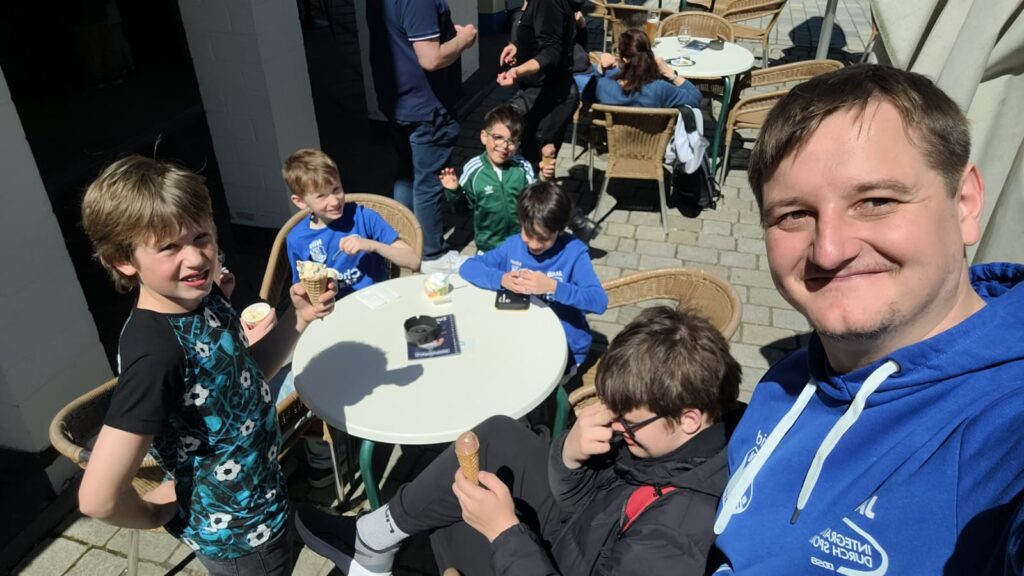 Man in a blue hoodie takes a selfie with a group of kids eating ice cream at an outdoor cafe table.
