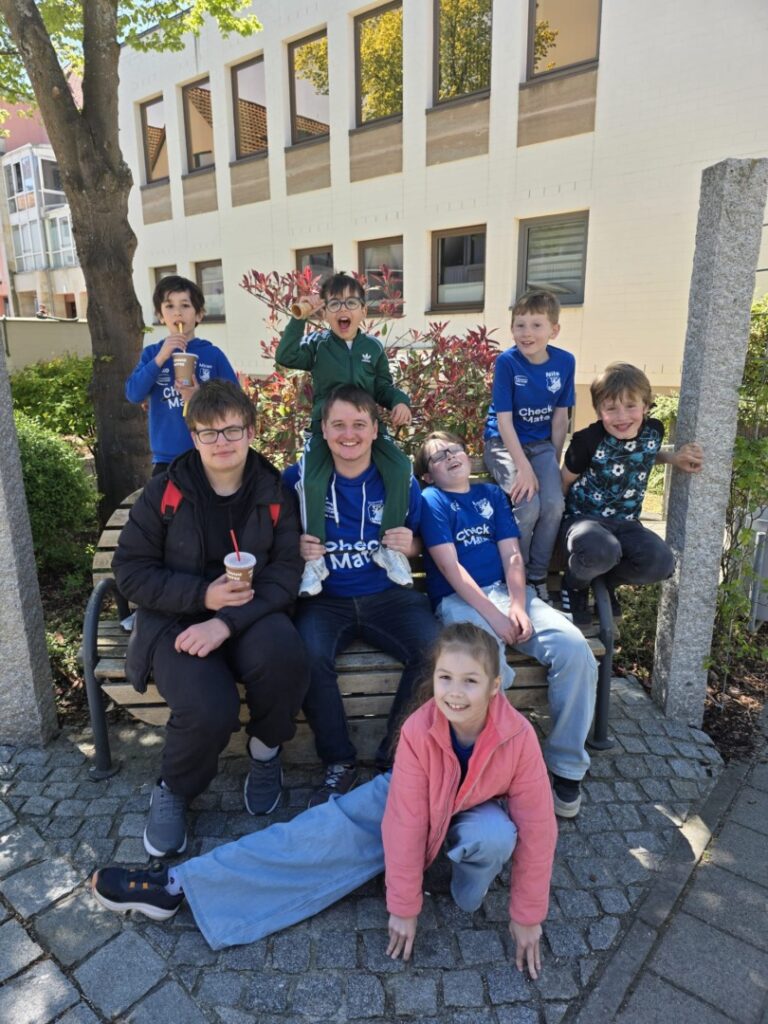 A group of kids wearing blue shirts sit on a bench outside a building, smiling for the photo with one child on a friend's shoulders and another lying on the ground in front.