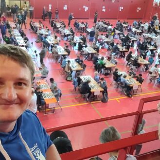 Smiling man in a blue hoodie takes a selfie from a balcony overlooking a gym filled with players at chess tables.