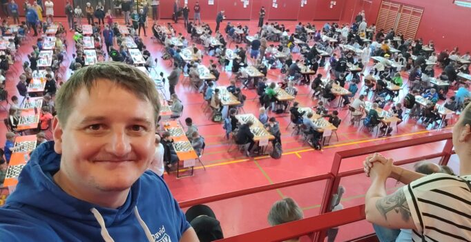 Smiling man in a blue hoodie takes a selfie from a balcony overlooking a gym filled with players at chess tables.