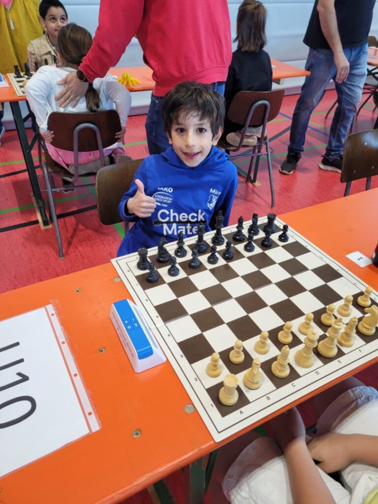 Smiling boy in a blue hoodie gives a thumbs up while seated at a chessboard ready for a match in a gym/classroom setting.