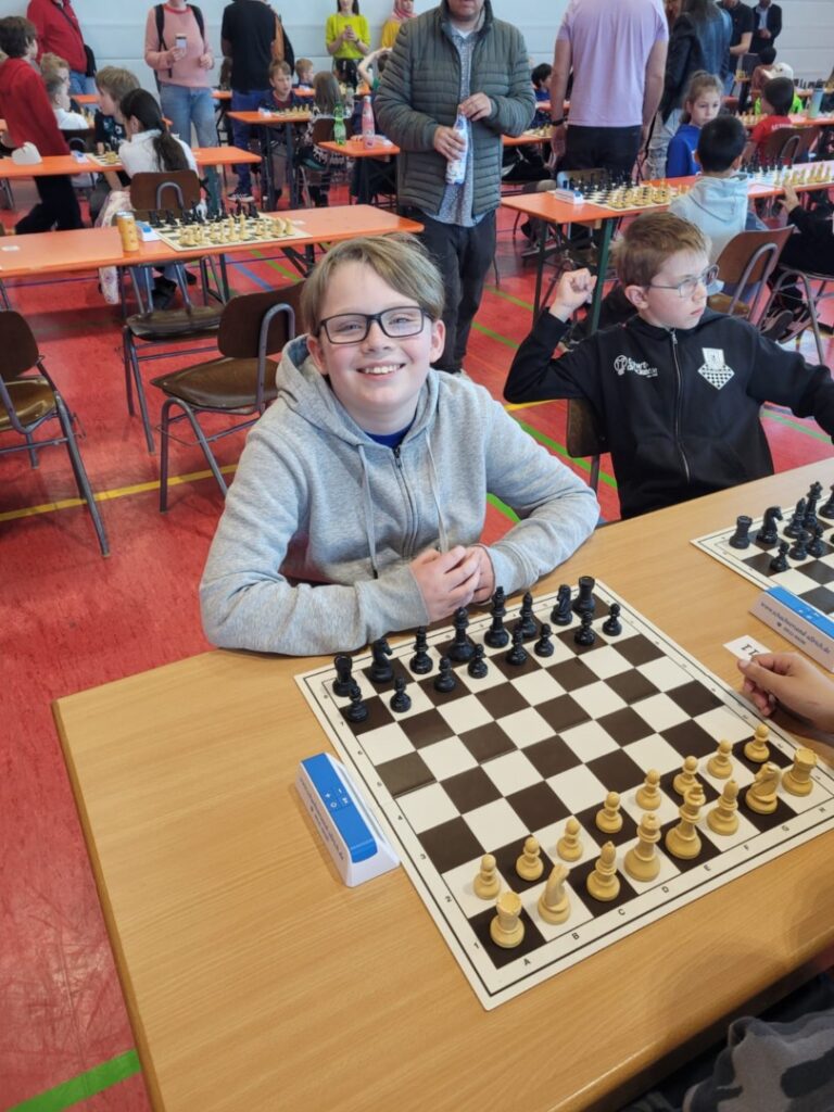 Smiling boy with glasses sits at a chessboard in a crowded gym during a youth chess tournament.