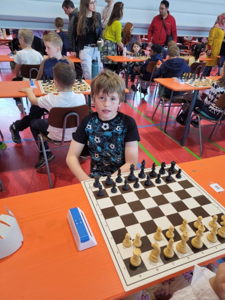 Young boy sits at an orange table playing chess at a crowded kids’ tournament hall with other players and spectators in the background.
