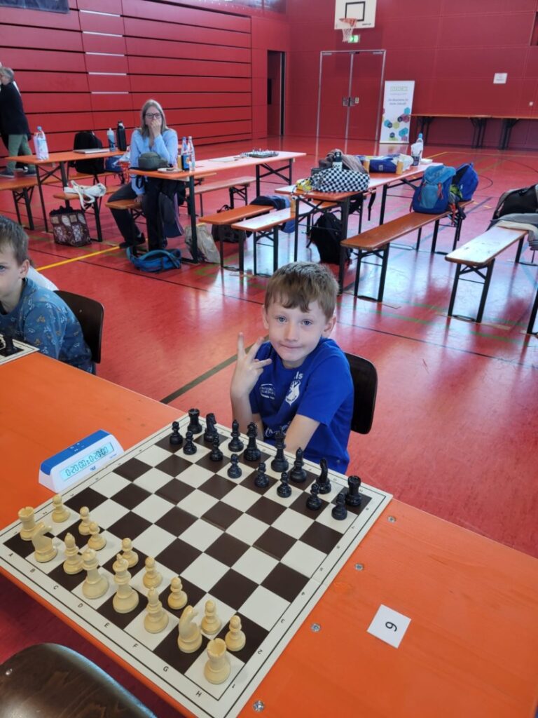 A young boy in a blue shirt sits at a chessboard in a gym, making a peace sign with his fingers; other players and bags are on benches behind him.