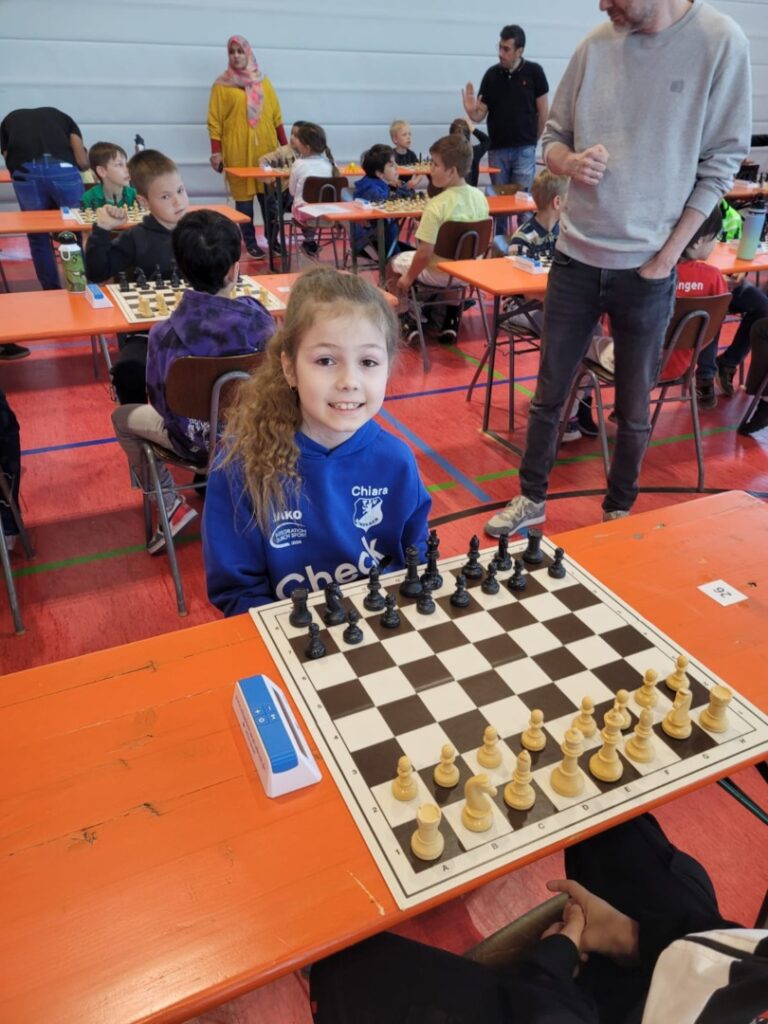 Young girl in a blue hoodie sits at a chessboard during a kids' tournament in a gym.