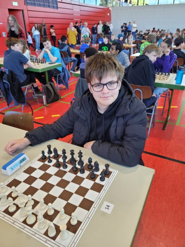 Young man with glasses sits at a chessboard during a tournament in a gym, with players at tables in the background.