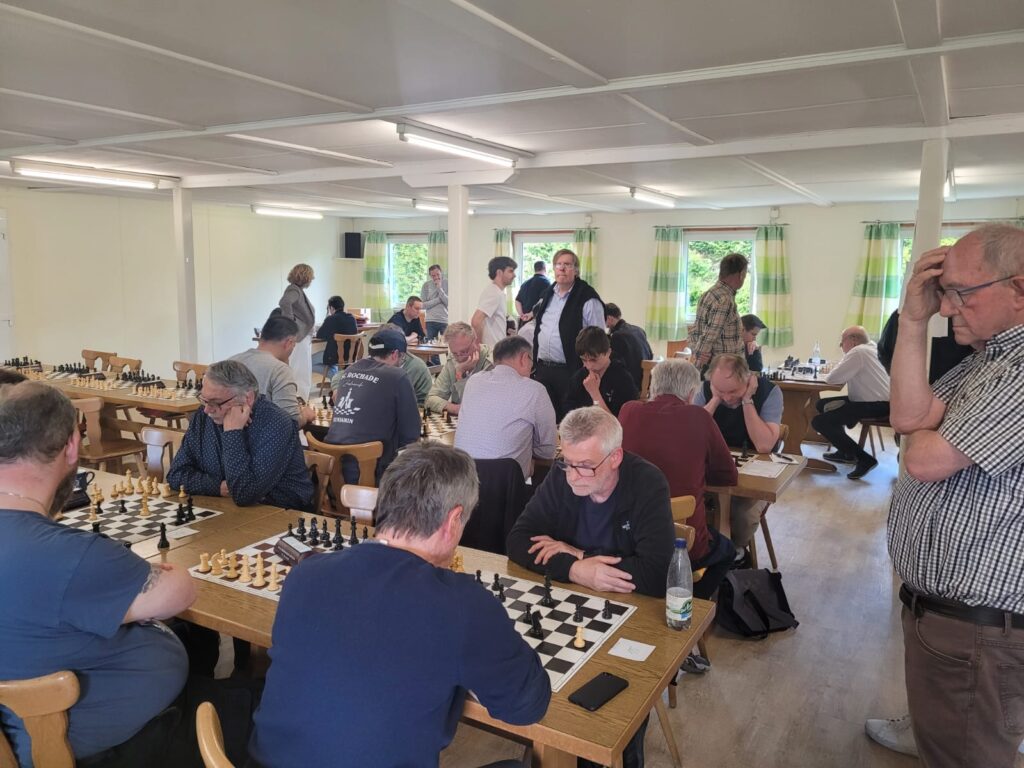 People playing multiple chess games at long wooden tables in a bright room with green-striped curtains.",
